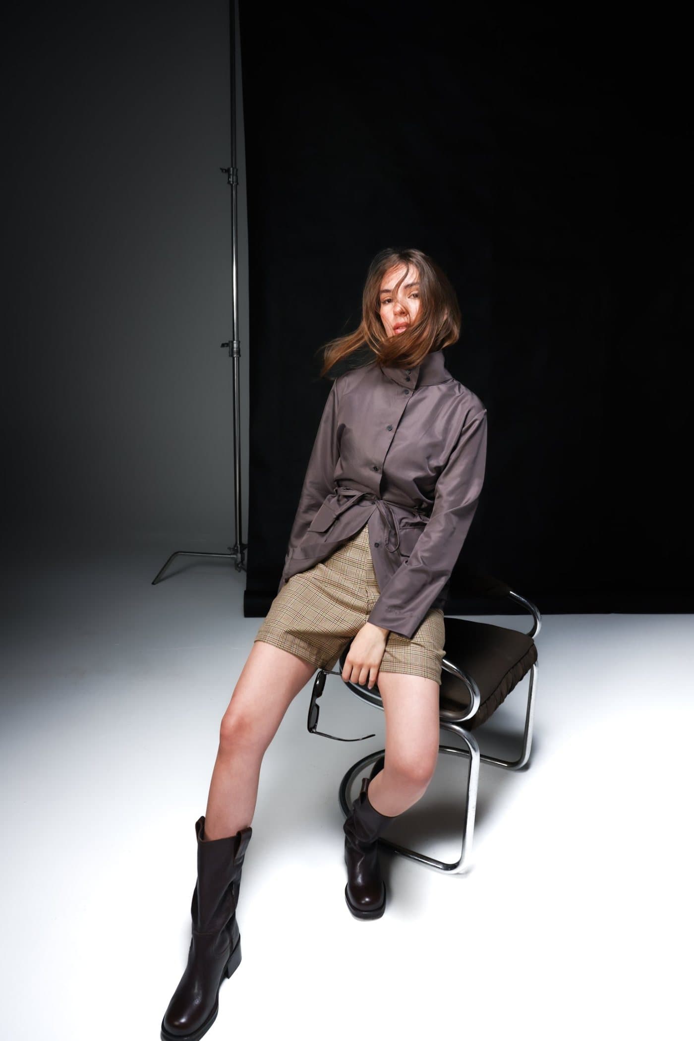 Model seated on a chrome chair in the chocolate shirt jacket and glen check short